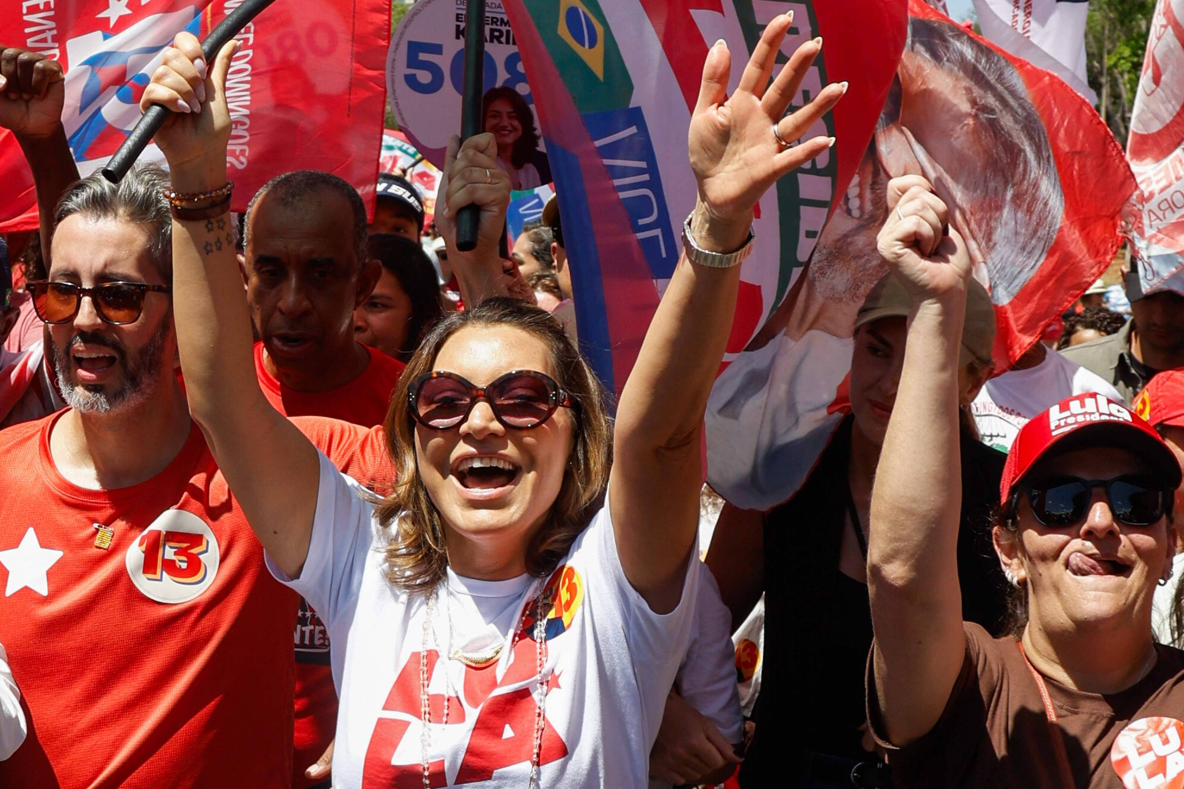 Grupo de pessoas em manifestação segurando bandeiras vermelhas e levantando os braços. Algumas vestem camisetas com o número 13 e símbolos políticos, incluindo uma mulher sorridente de óculos escuros no centro.
