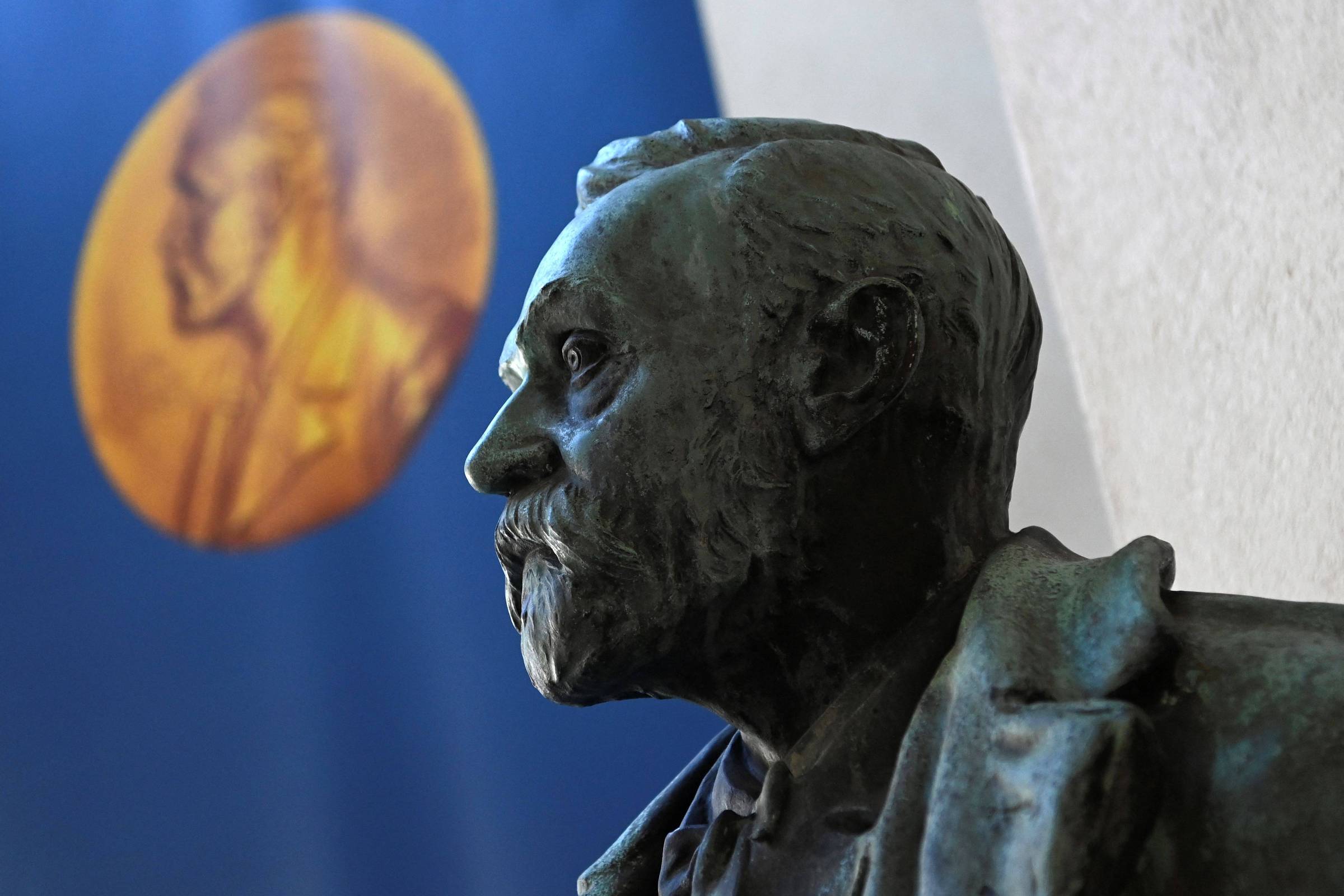 Busto em bronze de Alfred Nobel em perfil esquerdo, com detalhes realistas no rosto e barba. Ao fundo, desfocada, está uma grande medalha dourada com o perfil de Nobel, sobre um fundo azul.