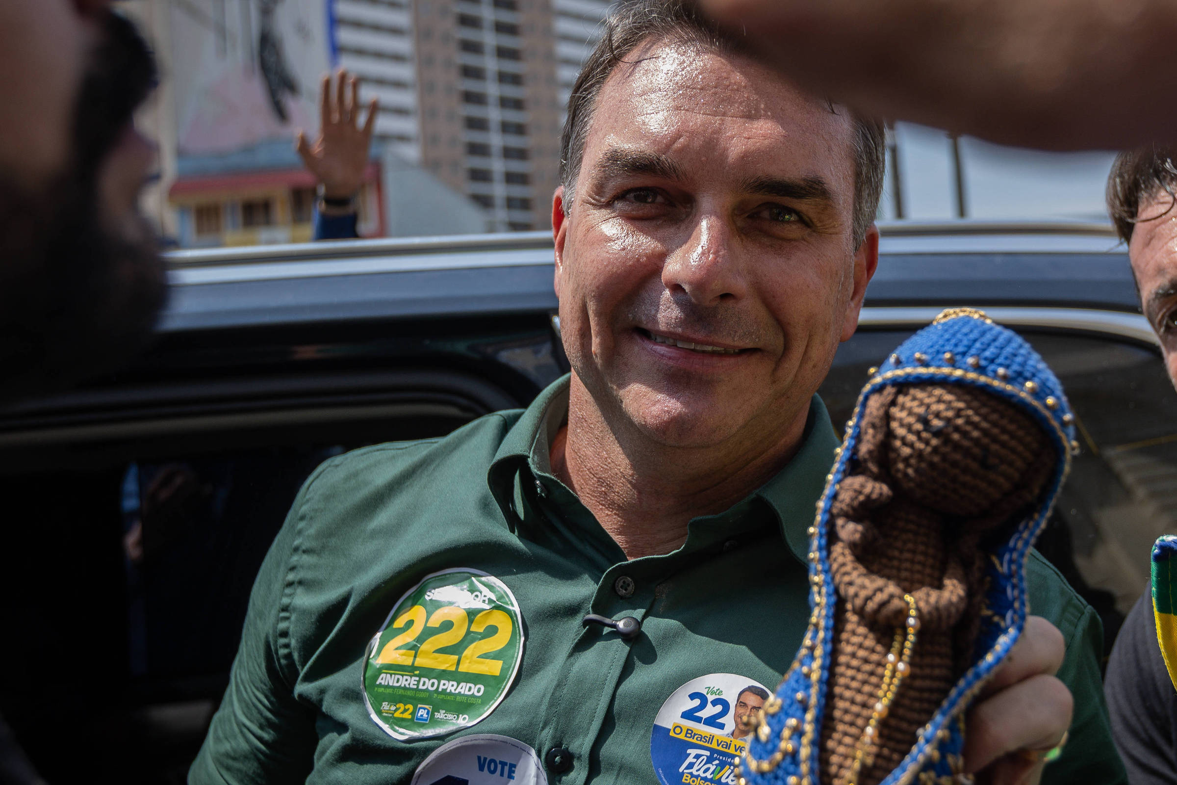 Homem com camisa verde e adesivos de campanha segura imagem de Nossa Senhora do Rosário em destaque. Outras três pessoas estão próximas, uma delas usa chapéu com as cores da bandeira do Brasil. Veículo e edifícios aparecem ao fundo.