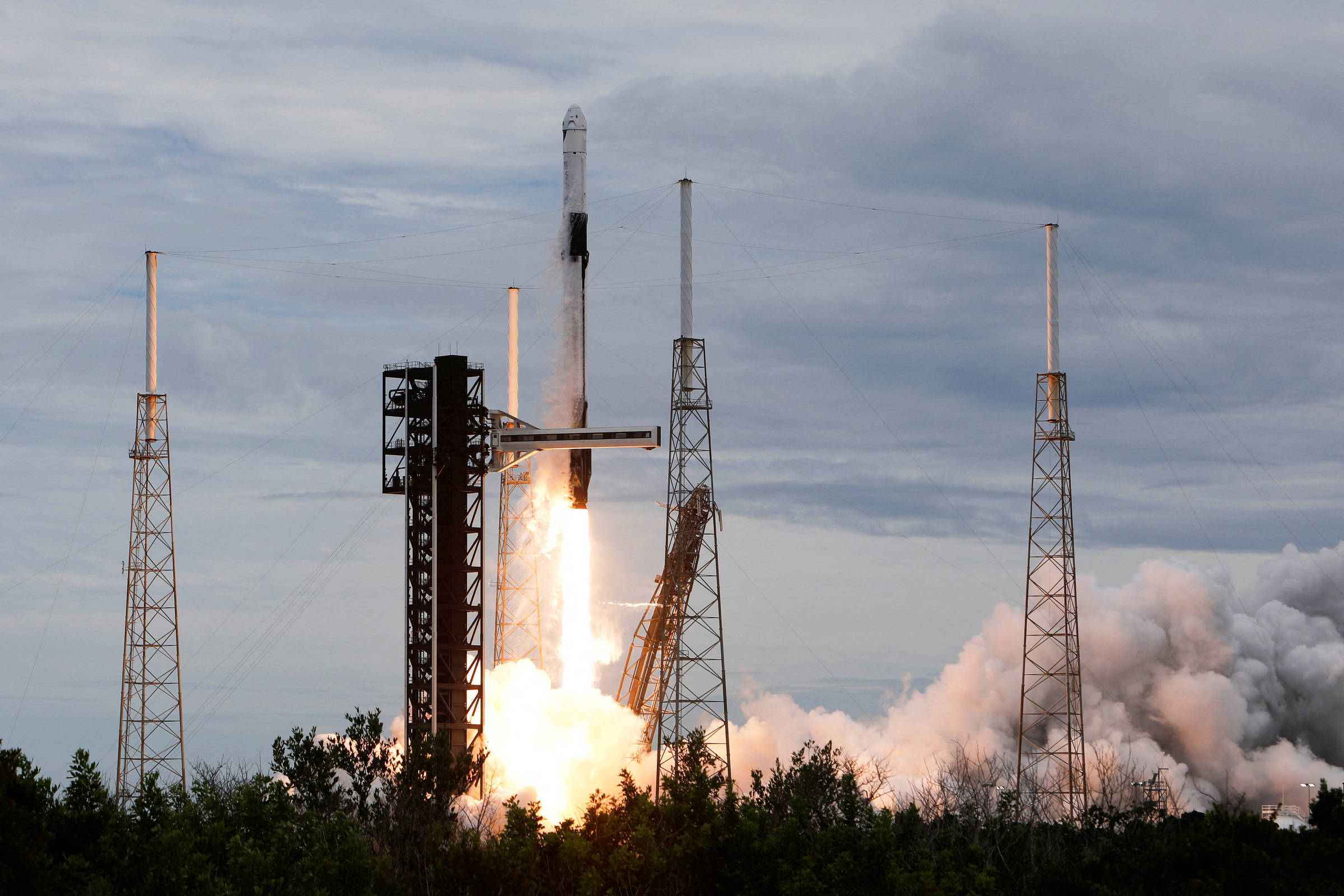 Foguete Falcon 9 da SpaceX decola da plataforma de lançamento com chamas e fumaça intensa na base. Estruturas metálicas altas cercam a plataforma sob céu nublado.