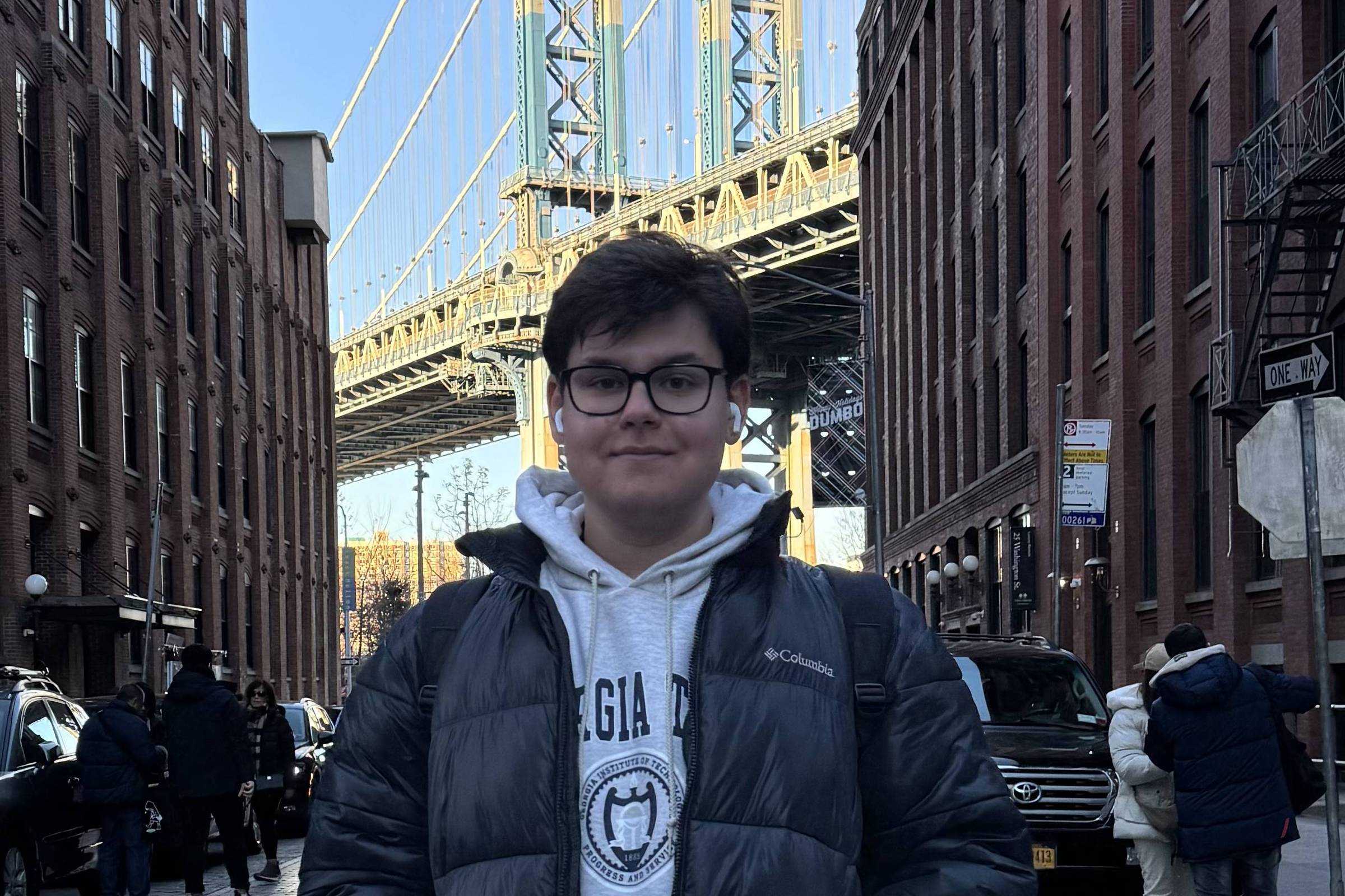 Jovem de óculos e jaqueta preta está em rua entre prédios de tijolos vermelhos, com a ponte Manhattan ao fundo sob céu azul.