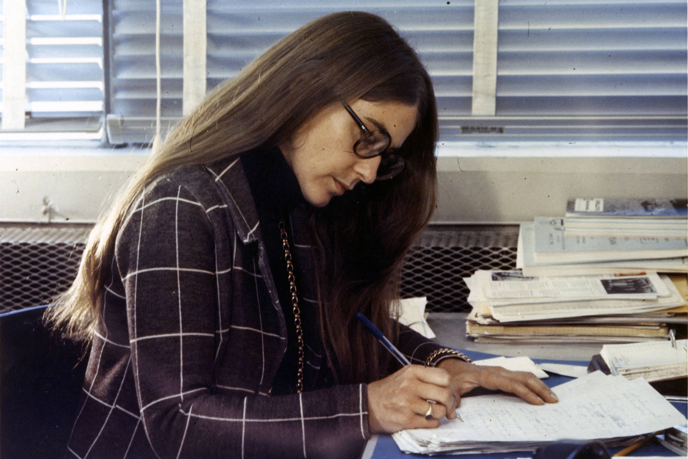 Mulher de cabelos longos e óculos está sentada à mesa escrevendo em caderno. Ela veste blazer xadrez escuro e está em ambiente com janela e pilhas de papéis ao fundo.