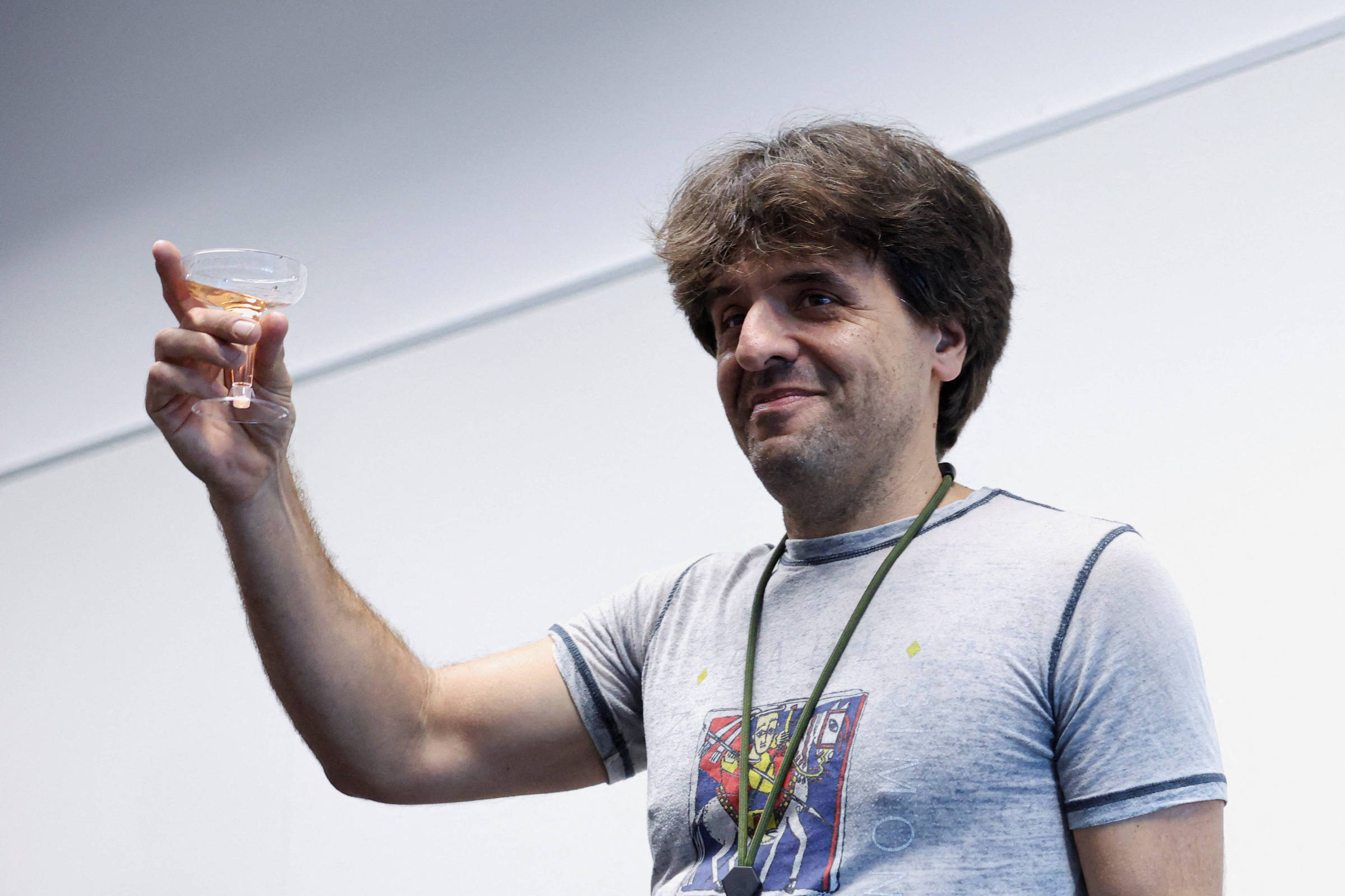 Homem de cabelos castanhos e camiseta cinza com estampa segura uma taça de vidro com líquido rosado, erguendo-a em gesto de brinde contra fundo branco.