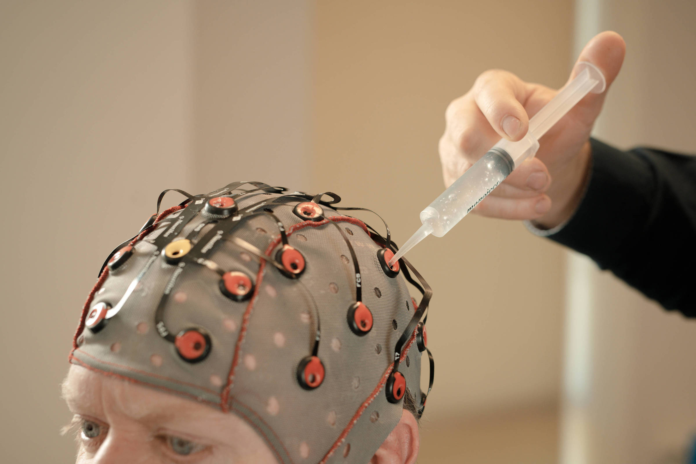 Pessoa usando capacete cinza com múltiplos eletrodos vermelhos conectados por fios pretos. Mão segura seringa próxima a um eletrodo no capacete.