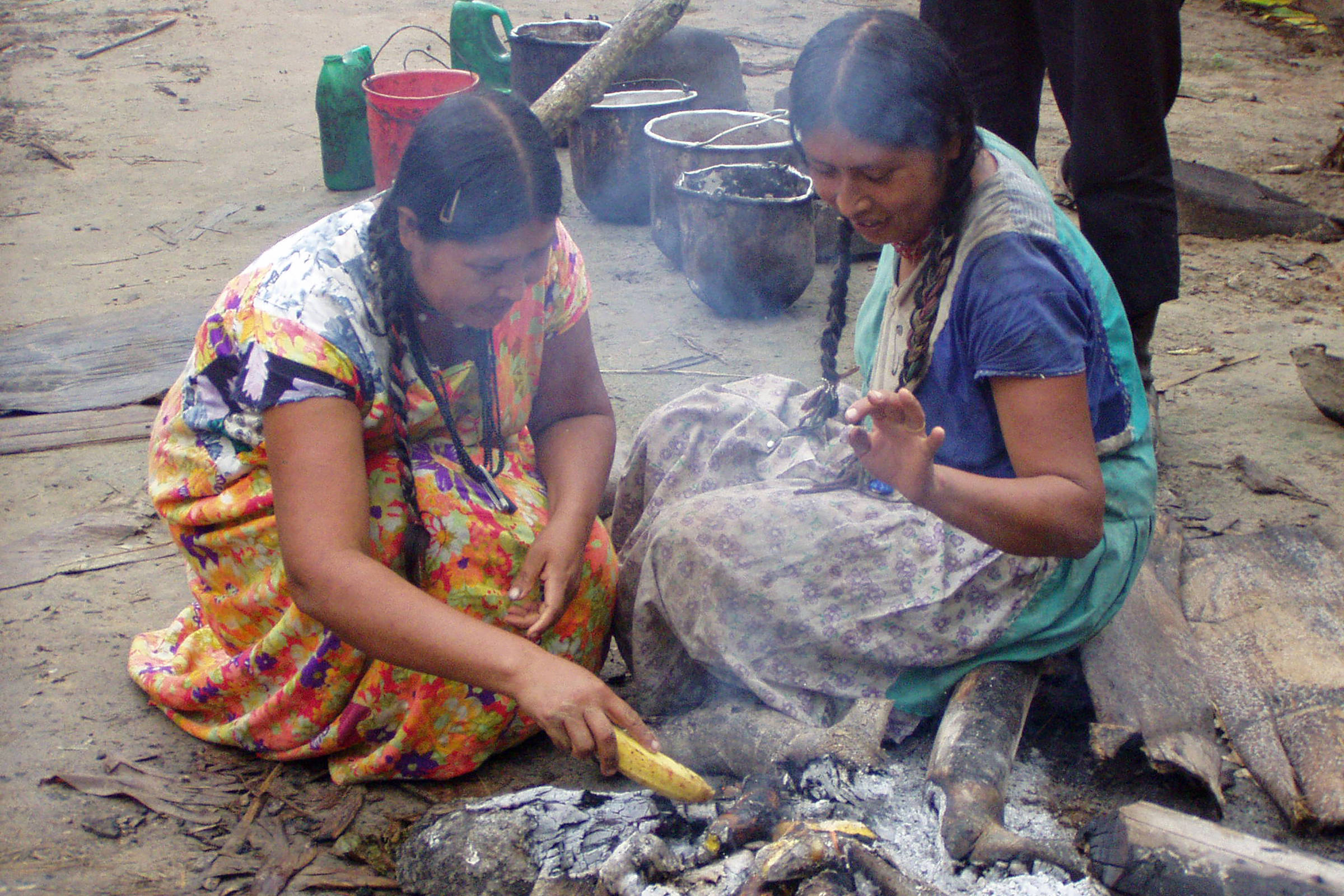 Duas mulheres indígenas sentadas no chão preparam alimentos em uma fogueira ao ar livre. Elas usam vestidos coloridos e têm cabelos trançados. Ao fundo, utensílios e recipientes estão dispostos sobre o solo.