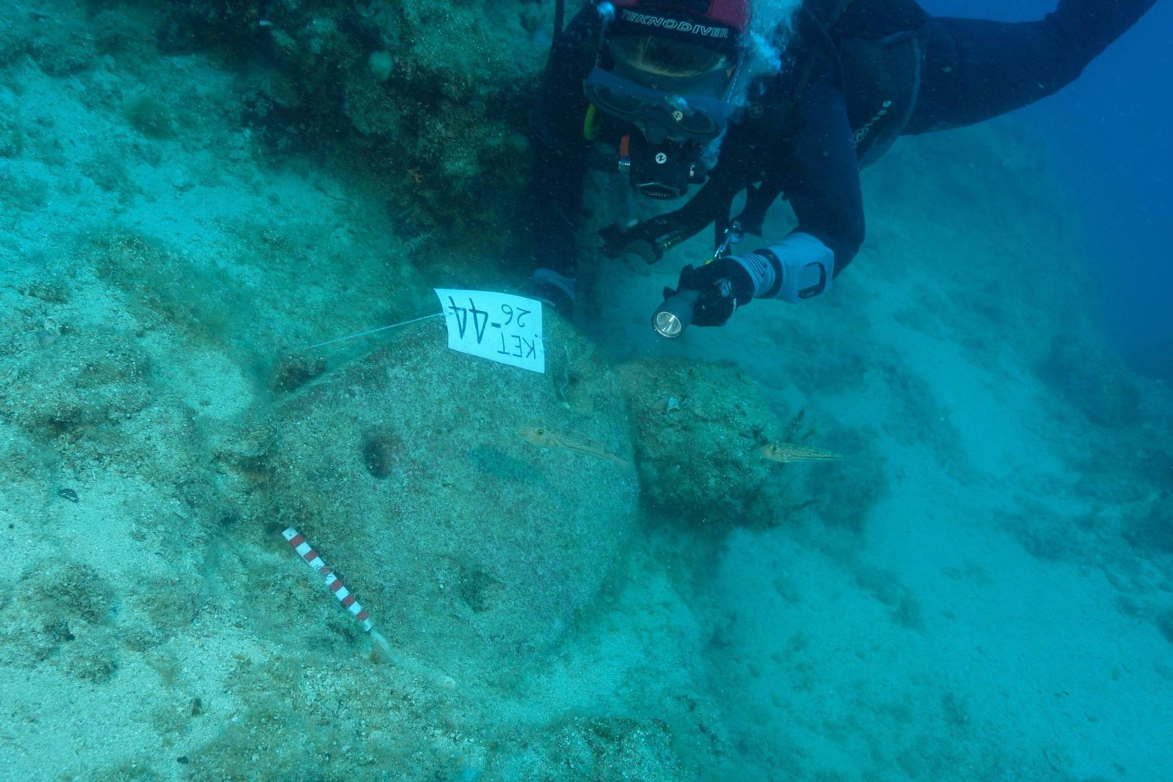 Mergulhador com equipamento completo registra com câmera artefato arqueológico no fundo do mar. Placa com código