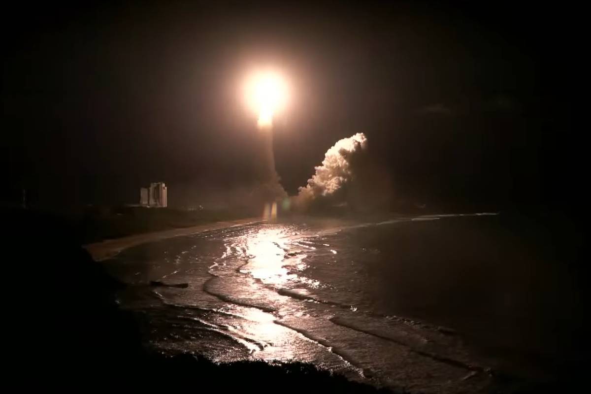 Foguete é lançado à noite de uma plataforma próxima à costa, iluminando o céu escuro. Reflexo da luz do foguete aparece na água abaixo, com fumaça visível ao redor da base.