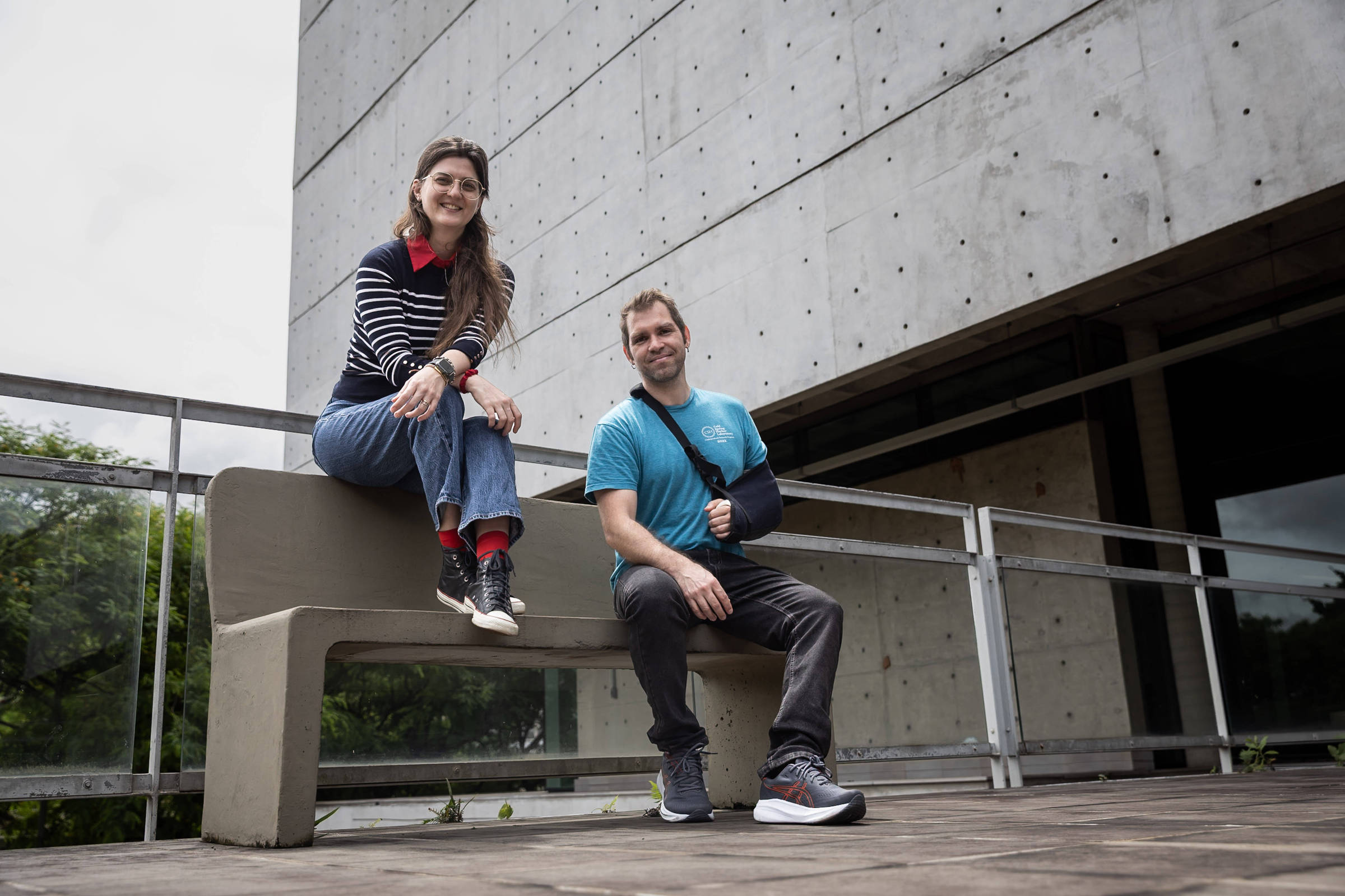 Uma mulher com blusa listrada preta e branca, calça jeans e tênis pretos está sentada no encosto de um banco de concreto. Ao lado dela, um homem com camiseta azul, calça preta e tênis cinza está sentado no assento do banco, com o braço direito imobilizado por tipoia preta. Ambos estão em área externa com piso de pedra, próximo a um prédio de concreto com janelas escuras e guarda-corpo metálico.