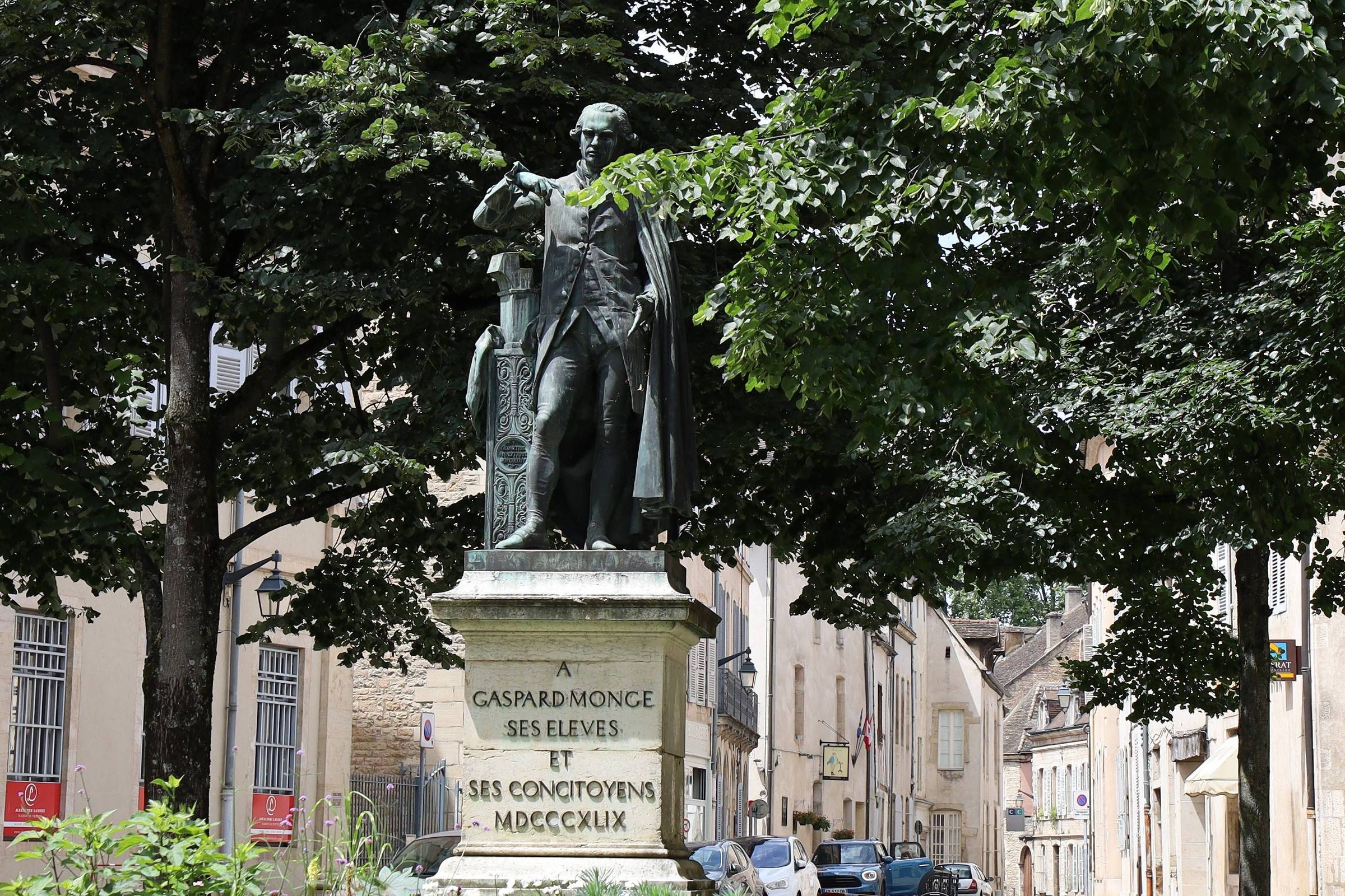 Estátua de bronze de Gaspard Monge em pedestal de pedra com inscrição, rodeada por árvores e edifícios históricos ao fundo em rua urbana.
