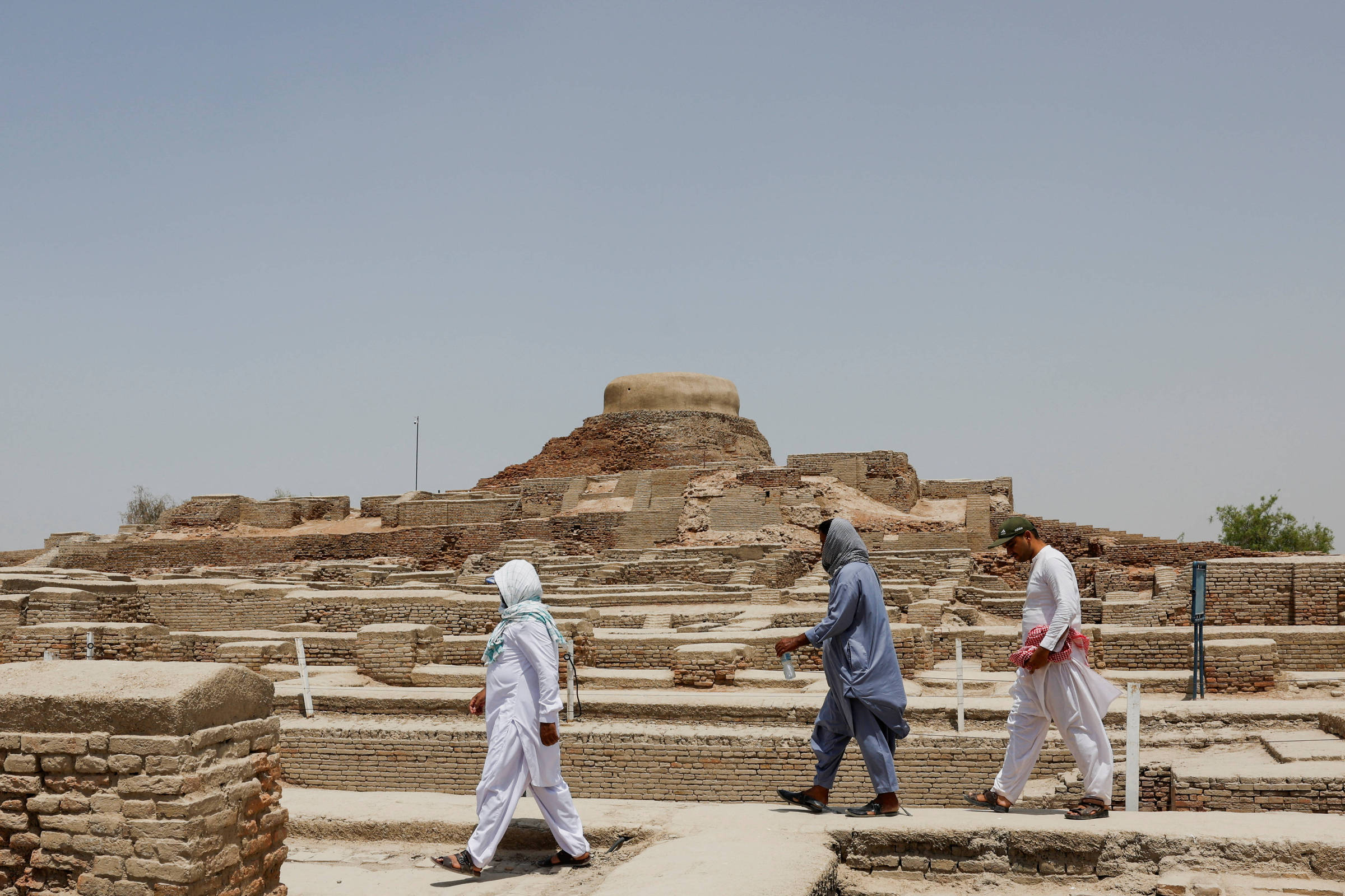Três pessoas vestindo roupas tradicionais caminham entre as ruínas de tijolos de Mohenjo-daro, sítio arqueológico no Paquistão, sob céu claro.