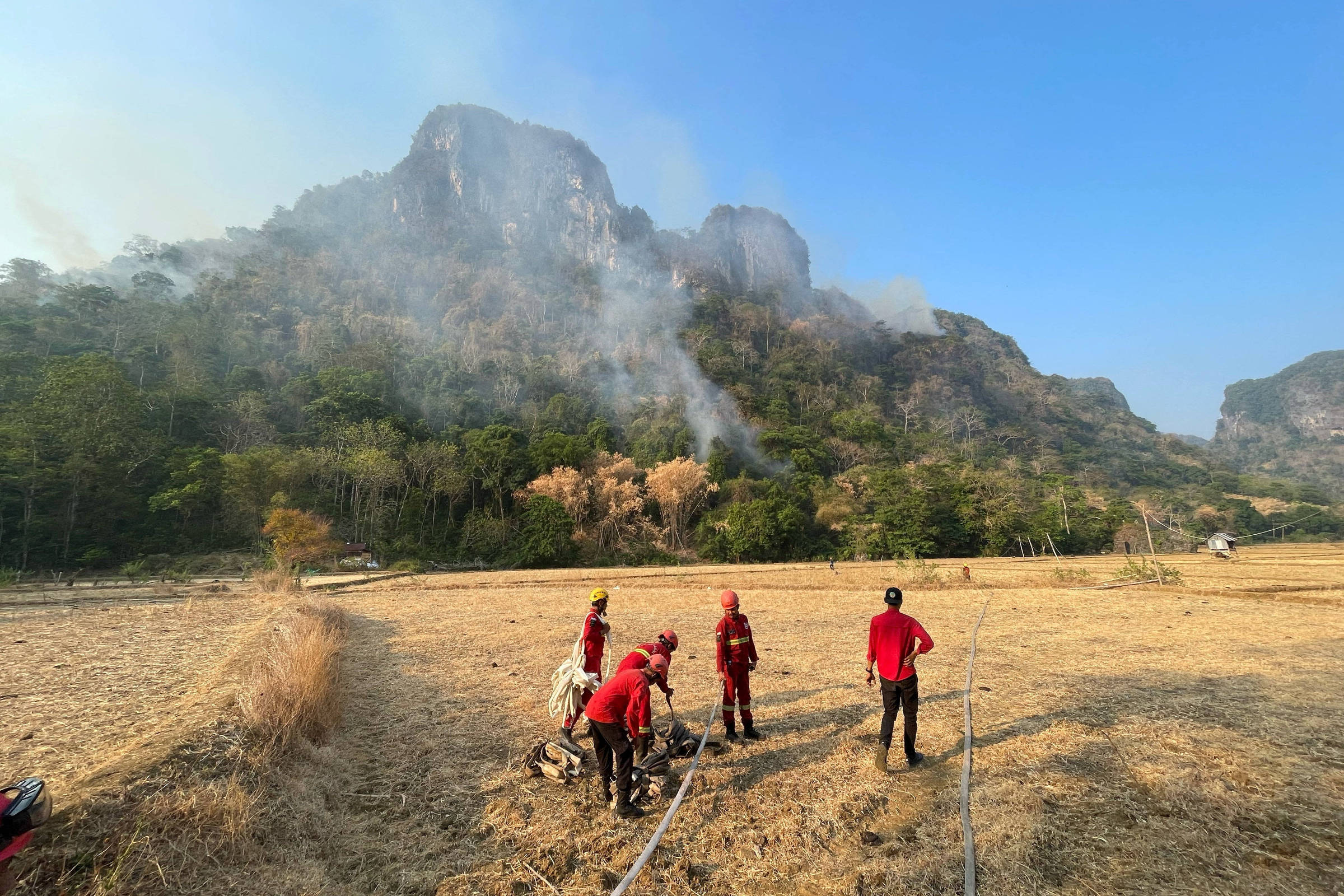 Cinco pessoas vestindo roupas vermelhas trabalham em campo seco próximo a uma área de vegetação com fumaça subindo. Montanha rochosa e céu azul compõem o cenário ao fundo.