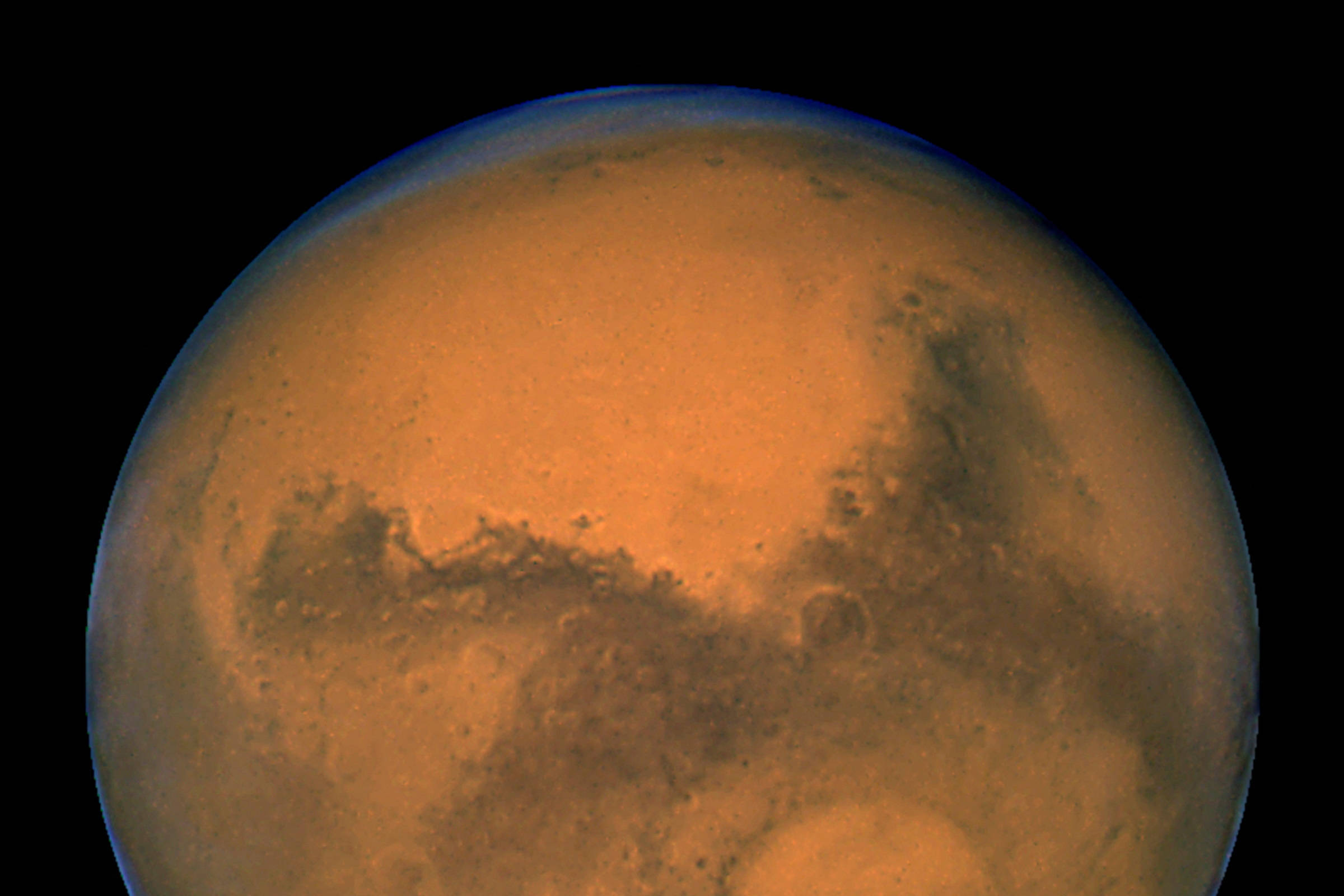 Imagem do planeta Marte mostrando sua superfície avermelhada com áreas escuras e uma calota polar branca na parte inferior.