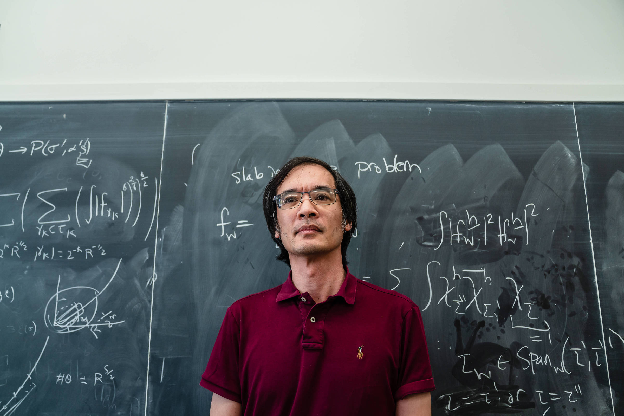 Homem de camiseta polo vinho está em pé diante de quadro negro coberto por fórmulas e equações matemáticas escritas com giz branco.
