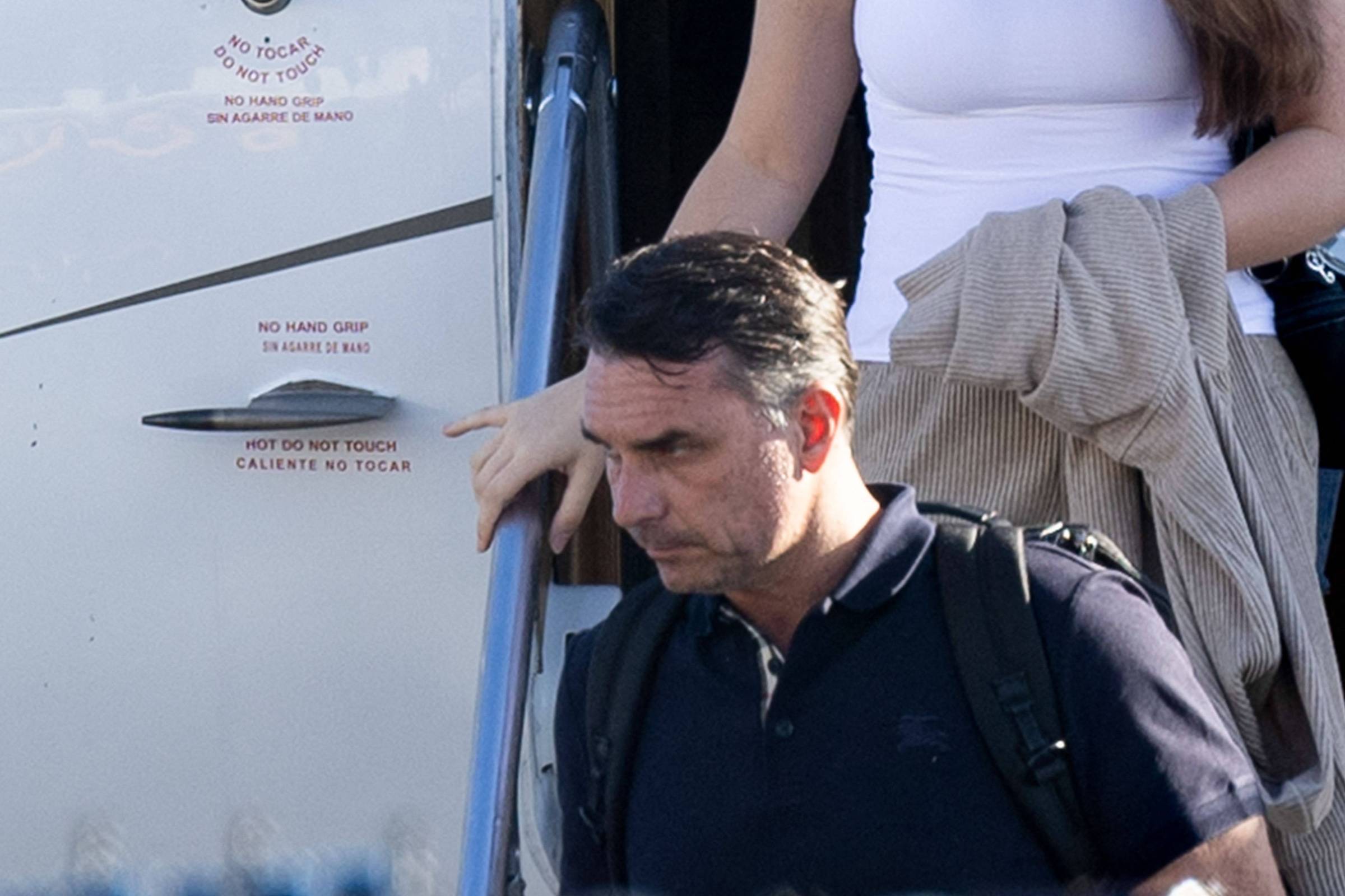 Homem com mochila preta desce escada de avião segurando corrimão. Mulher atrás veste regata branca e carrega casaco bege. Avisos em inglês e espanhol indicam para não tocar ou segurar corrimão.