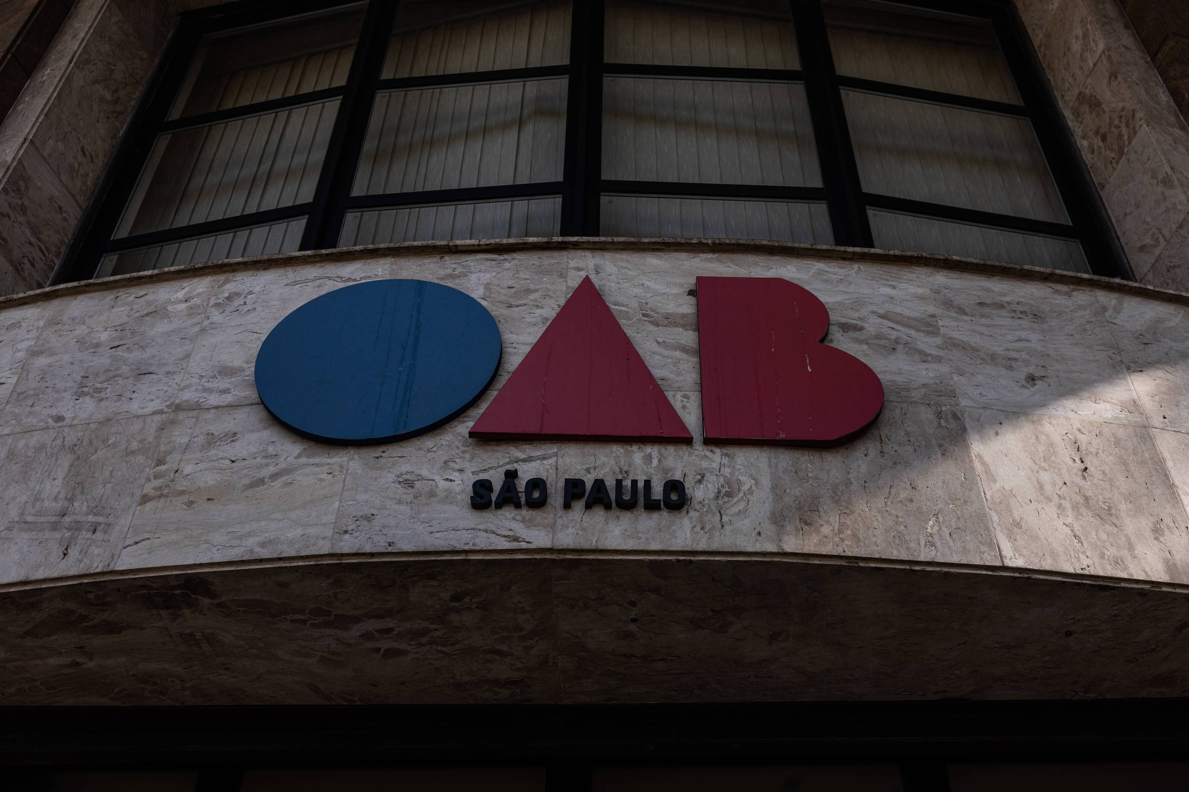 Logotipo da OAB em azul e vermelho fixado em fachada de pedra clara com janelas grandes ao fundo. Texto