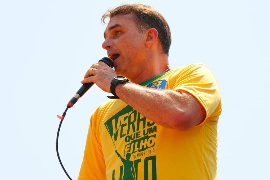 Homem de camiseta amarela com texto verde segura microfone e fala em evento ao ar livre sob céu claro.