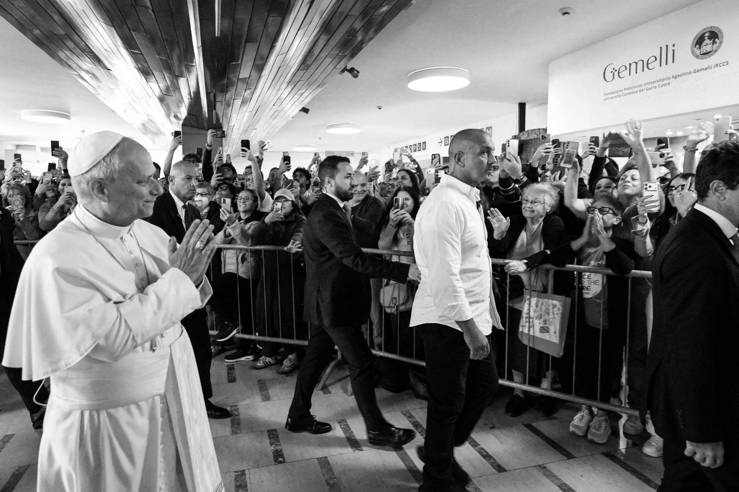 Papa Francisco caminha pelo corredor de hospital com mãos juntas em gesto de oração, enquanto multidão atrás de grades fotografa e acena. Segurança acompanha próximo a ele.