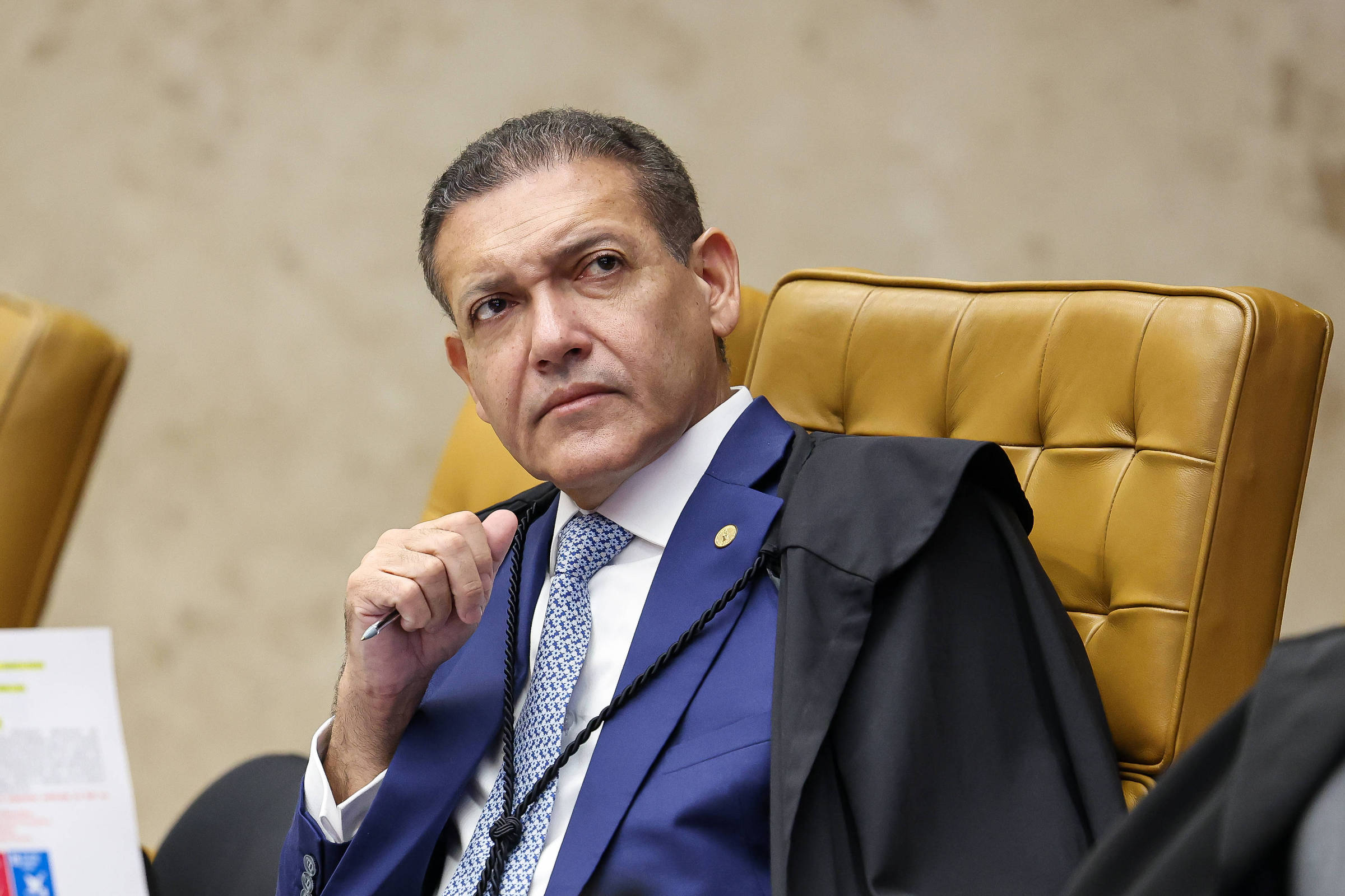 Homem de meia-idade com terno azul e gravata azul clara sentado em cadeira de couro marrom claro, segurando caneta próxima ao rosto, com expressão pensativa. Ao fundo, parede bege e parte de outra cadeira semelhante.