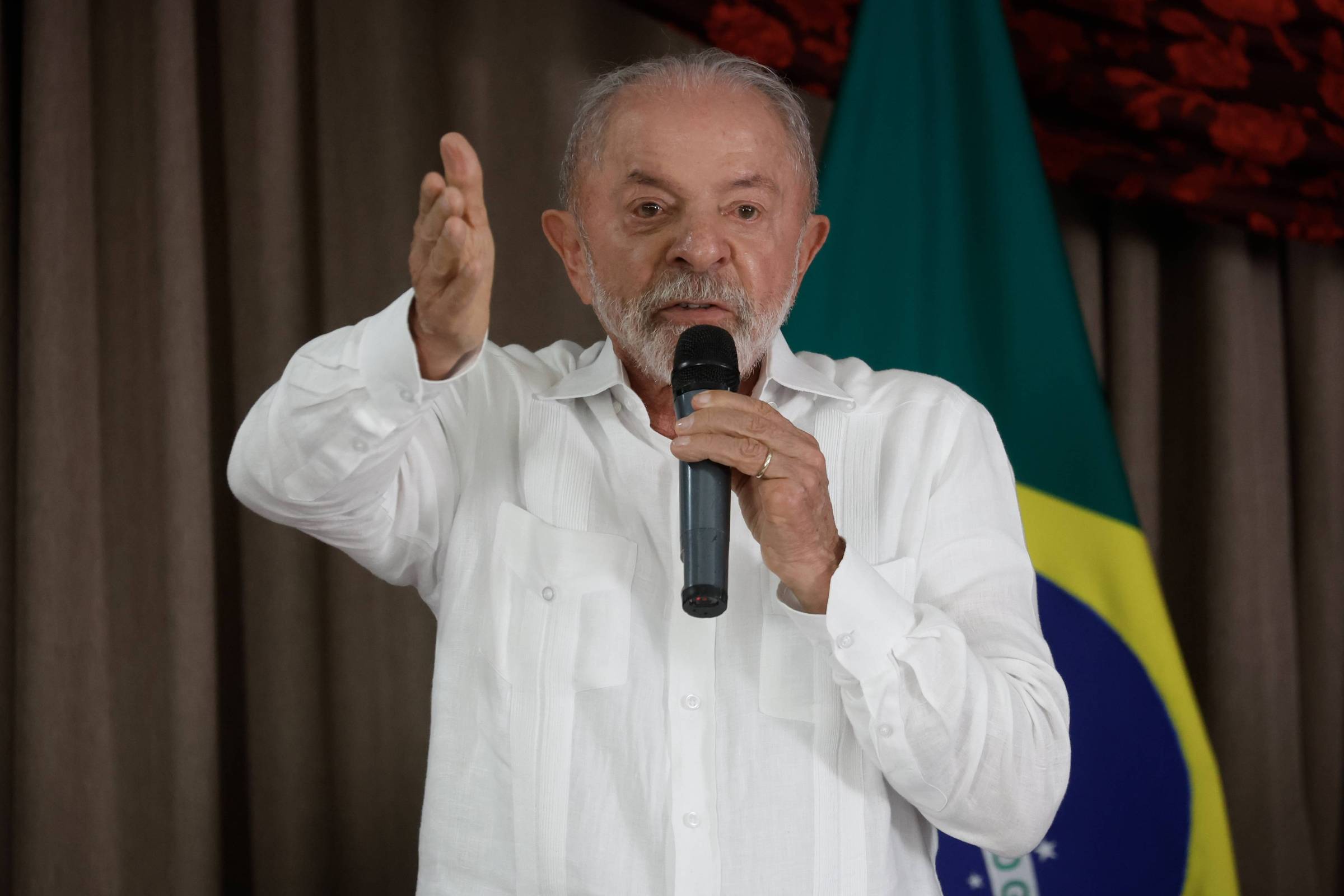 Homem de cabelos grisalhos e barba branca veste camisa branca e segura microfone com a mão direita enquanto gesticula com a esquerda. Ao fundo, bandeira do Brasil parcialmente visível contra cortina marrom.