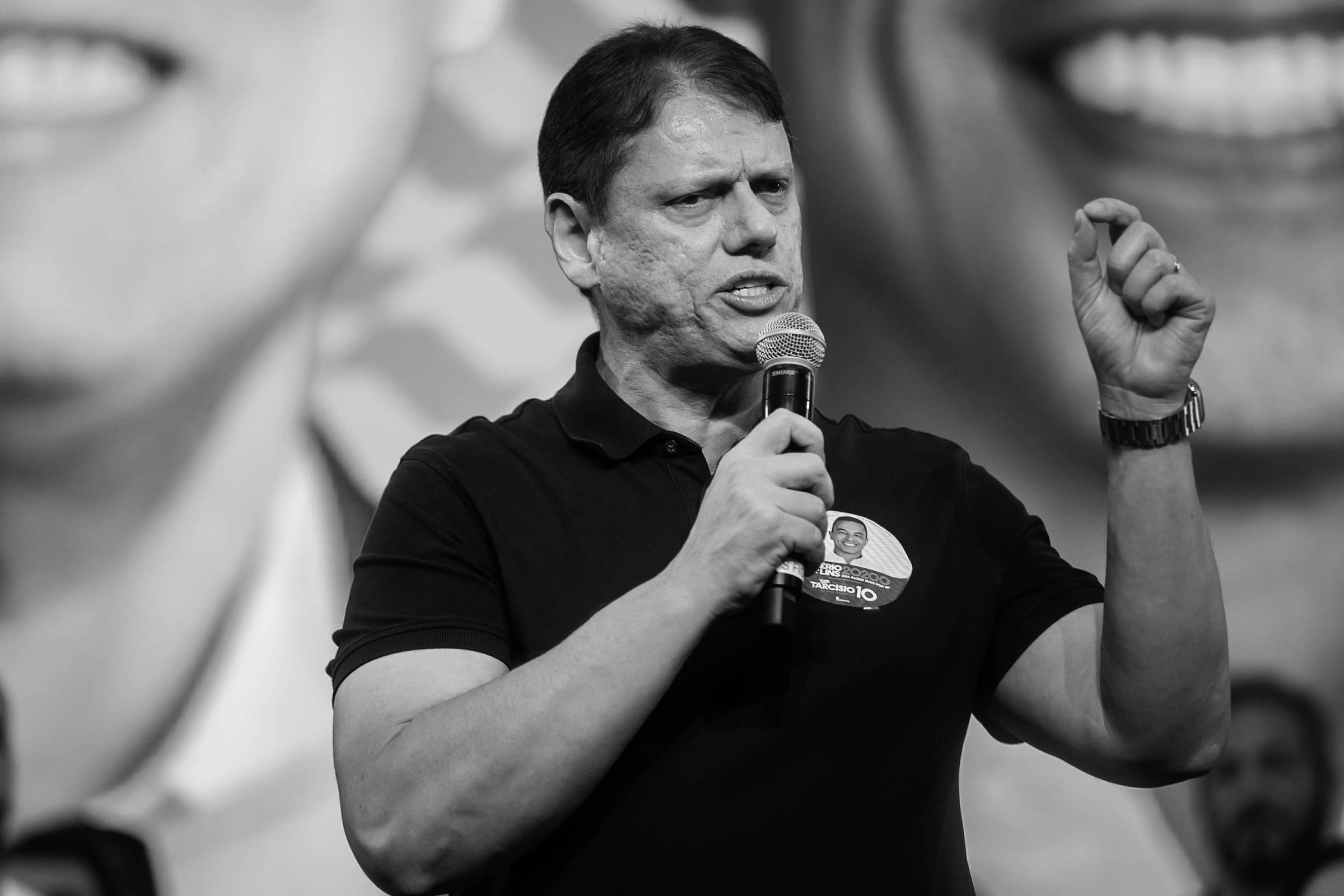 Homem de camiseta preta segura microfone e gesticula durante discurso em evento. Fundo exibe imagem ampliada de rosto sorridente.